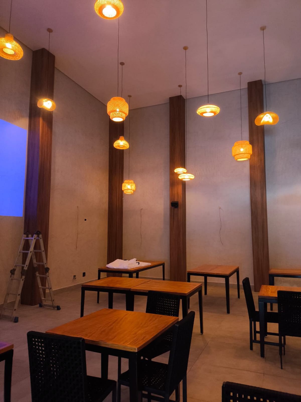 Salão de restaurante com parede em gesso e pé-direito alto, com luminárias pendentes entre pilares de madeira